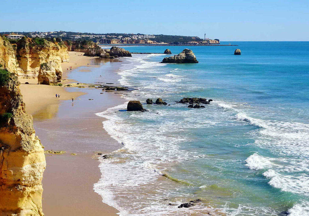 Waves crash onto a sandy beach lined with golden cliffs and scattered rock formations along the shoreline.