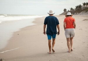 Older couple walking barefoot along a sandy beach, both wearing sun hats and casual summer clothes.