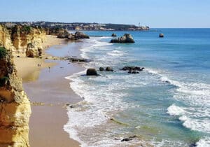 Waves crash onto a sandy beach lined with golden cliffs and scattered rock formations along the shoreline.