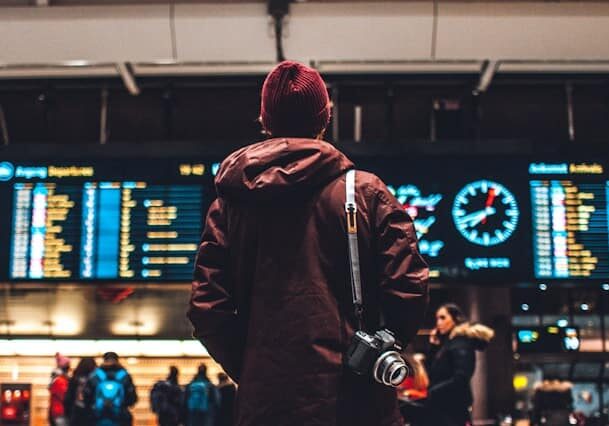 Back view of a man in a red beanie standing in front of an arrivals board at an airport - second passport received through investment