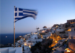 A coastal village in Greece at night, illuminated by warm yellow lights, with the Greek flag waving in the foreground.