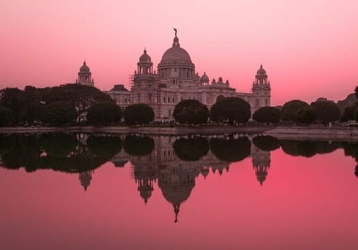 Historic building with domes and towers reflecting in a calm lake under a pink-hued sky at sunset.