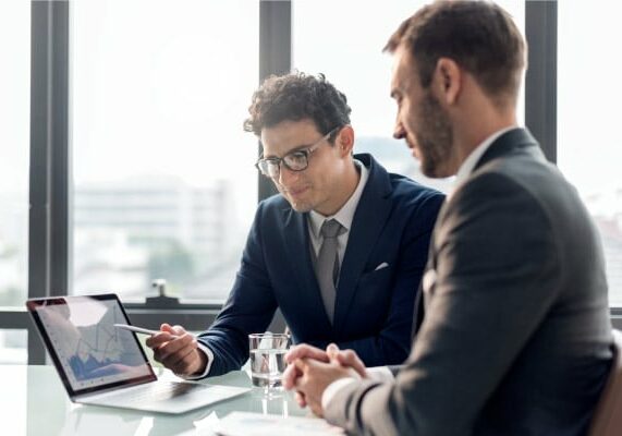 Two businessmen in suits discussing a financial chart on a laptop in a modern office with large windows.