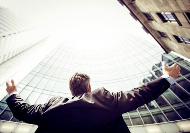 Businessman standing with arms raised, looking up at tall glass buildings in a cityscape.