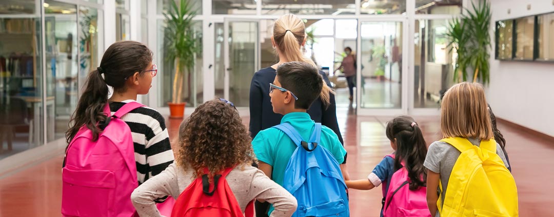 Children with backpacks following a teacher. 
