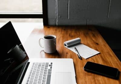 laptop on a desk with a phone, notebook, pen and coffee