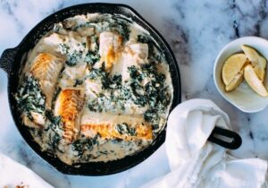 Salmon fillets in creamy spinach sauce cooking in a black skillet on a marble countertop.
