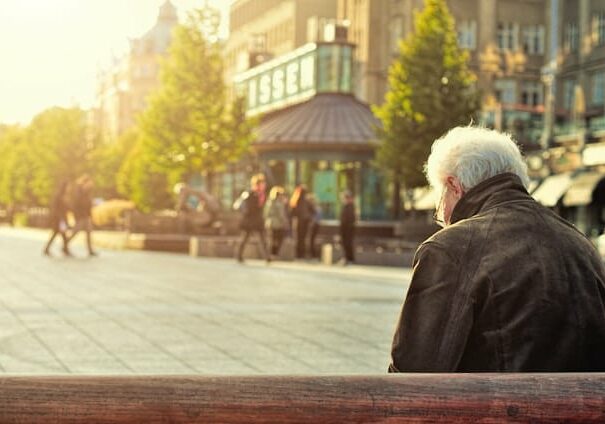 Back view of man with gray hair sitting on a bench in a town square with golden light surrounding him - where to retire in Europe