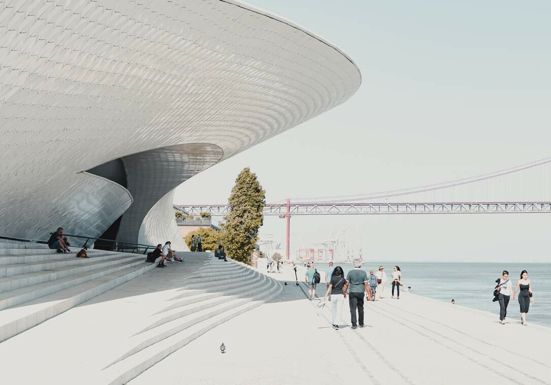 People walking along the waterfront near the MAAT museum in Lisbon, with the 25 de Abril Bridge in the background.