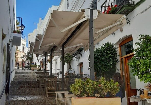 Two white buildings opposite each other in Spain, with a restaurant featuring chairs and tables outside, and a walkway with stairs in between.