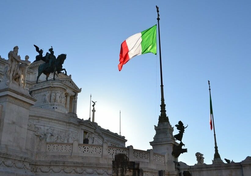 An Italian flag waving next to a monument with statues. 