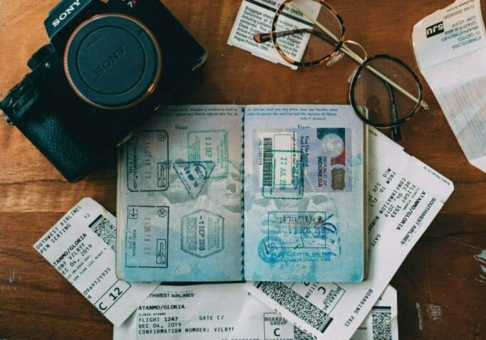 Open passport with various visa stamps surrounded by airline boarding passes, a Sony camera, and eyeglasses on a wooden table - how to buy a passport