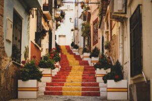 Stairs painted in Spanish flag colors with flowers on the side in a narrow street. 