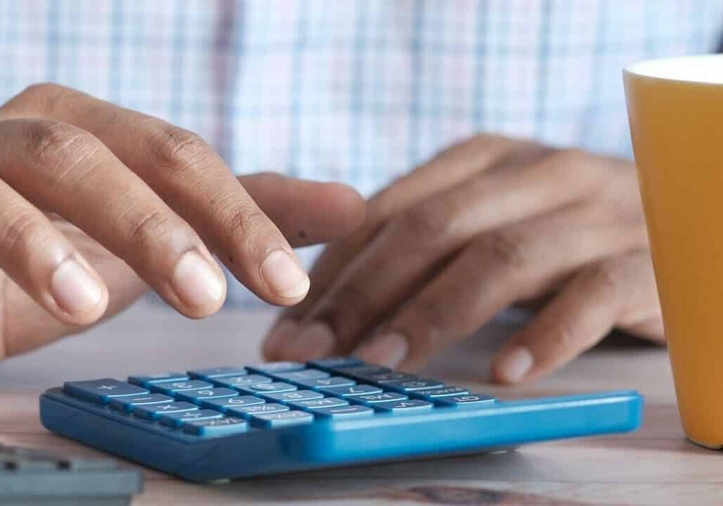 Close-up of hands using a blue calculator on a desk with an orange cup in the background.