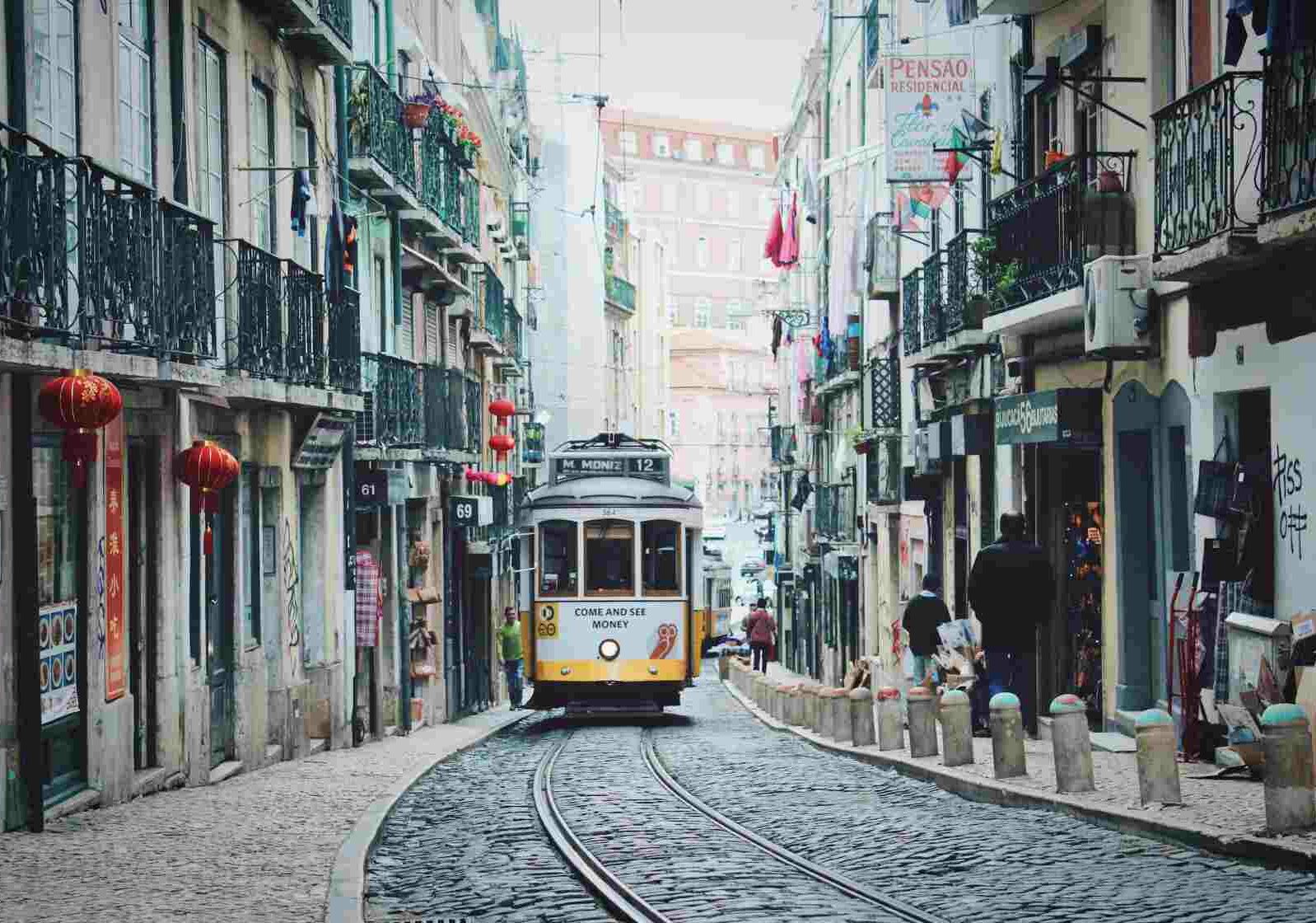 Historic yellow tram moving through a narrow, cobblestone street lined with old buildings and balconies in Lisbon.