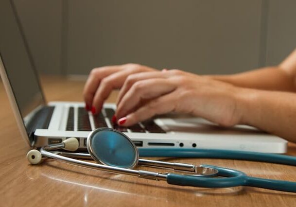 A person typing on a laptop with a stethoscope next to it. 