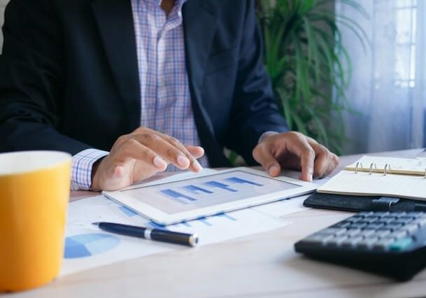 Businessman analyzing financial charts on a tablet at a desk with documents, calculator, pen, and coffee.