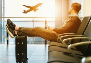 Man sitting in an airport terminal with legs on a suitcase, watching a plane take off through the window.