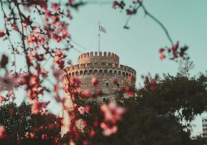 The White Tower of Thessaloniki framed by blooming pink flowers and trees under a clear sky with a Greek flag atop.