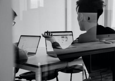 Black and white view through an office window of two people sitting in front of laptops talking and working - deducting business expenses as a means to legally avoid taxes