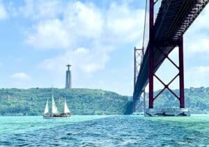 Sail boat under a bridge spanning over a body of water in Lisbon, Portugal - Foreigners who want to invest in Portugal for citizenship
