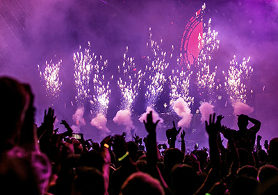 Vibrant music festival scene with a cheering crowd, colorful fireworks, and purple stage lighting during a live concert performance.