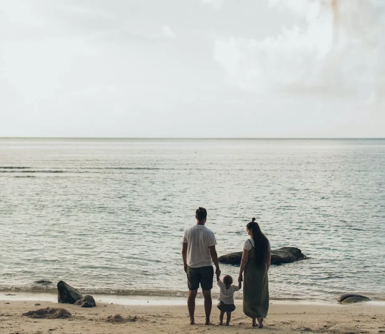 A family strolls along the beach, holding hands with their child, enjoying a sunny day by the ocean.