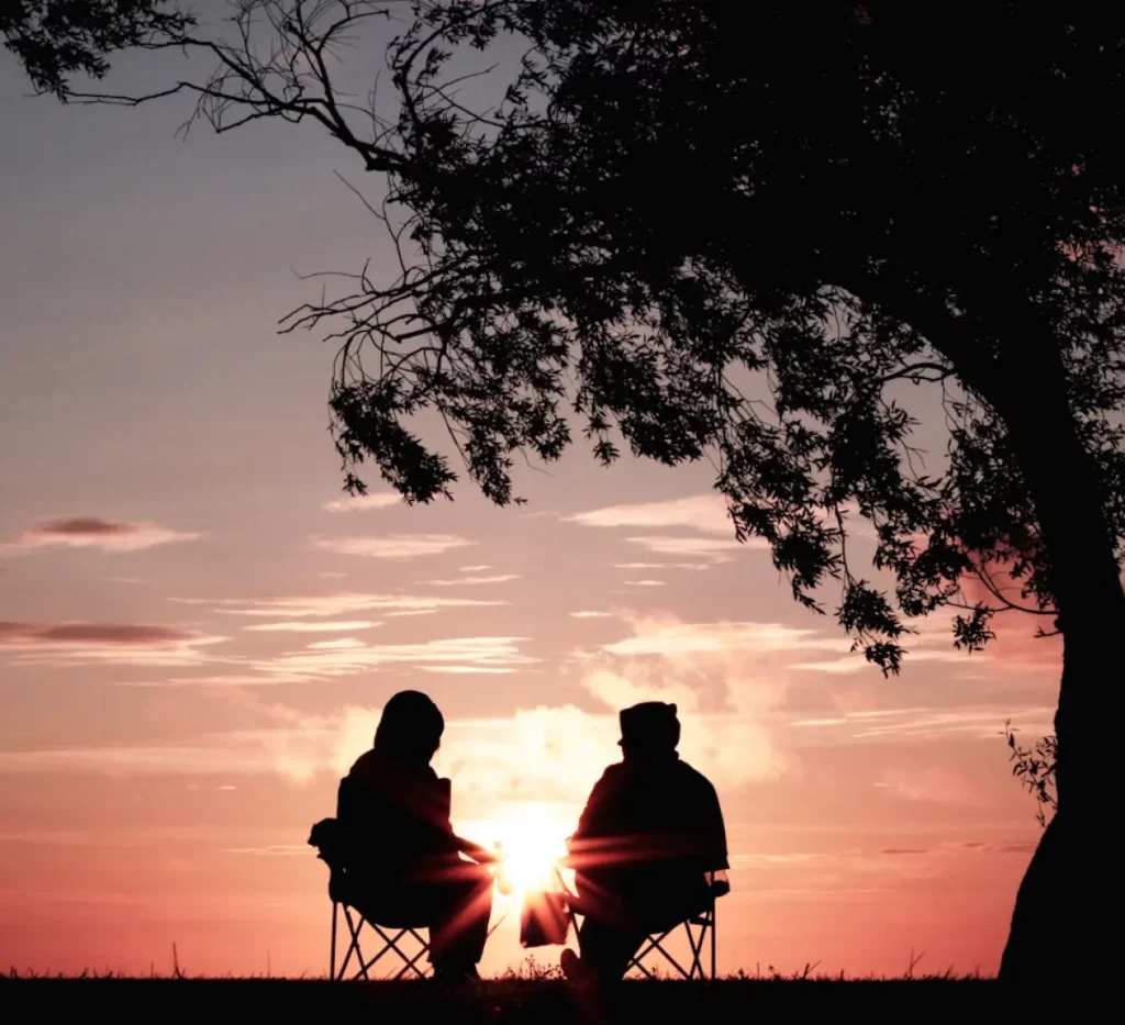 Two people sit in chairs under a tree, enjoying a serene sunset with warm colors illuminating the sky.