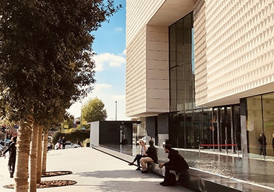 Modern architectural building in Istanbul with glass façades, people sitting near a shallow reflecting pool, and tree-lined pavement under a sunny sky.