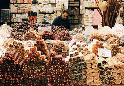 Colorful display of Turkish delight and sweets stacked in a market stall, with a vendor in the background surrounded by boxes and traditional treats.