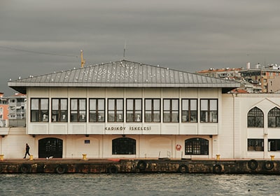 Kadıköy ferry terminal building in Istanbul, Turkey, viewed from the waterfront under a cloudy sky, with a pedestrian walking along the dock.