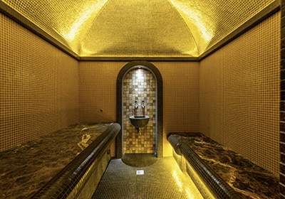 Luxurious Turkish hammam interior with mosaic-tiled walls, marble benches, and ambient golden lighting surrounding a central water fountain.