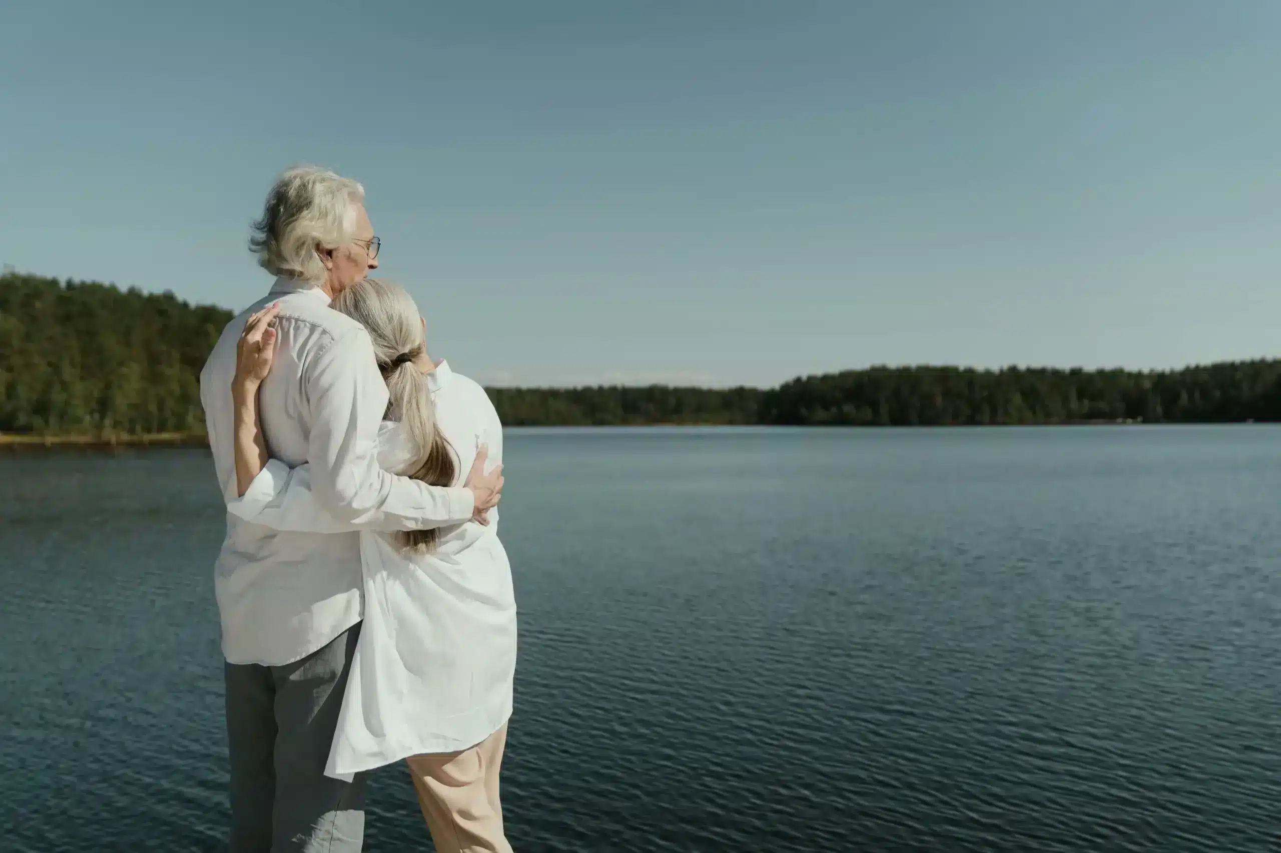 An older couple stands together by a serene lake, enjoying the peaceful scenery and each other's company.