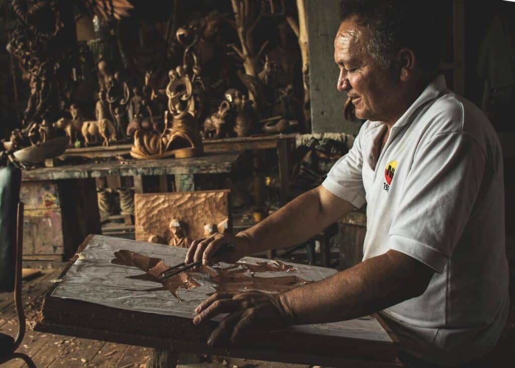 A man carves a wooden sculpture in his workshop, surrounded by tools and wood shavings.