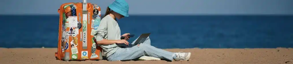 A person sitting on the beach, working on a laptop, with waves and sand in the background.