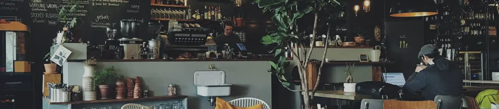 A man sits at a table in a coffee shop, focused on his laptop with a cup of coffee beside him.